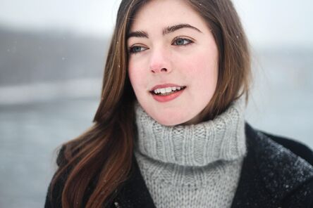 Junge Frau mit hellbraunen Haaren lächelt leicht und trägt einen warmen grauen Pullover bei Schneefall